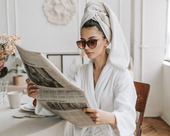 Woman in bathrobe reading a newspaper at a stylish indoor setting with flowers.