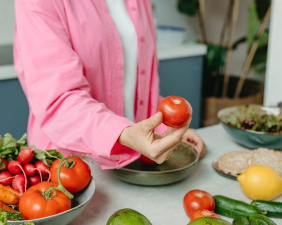 Person in pink overshirt holding a fresh tomato with various organic vegetables on a countertop.