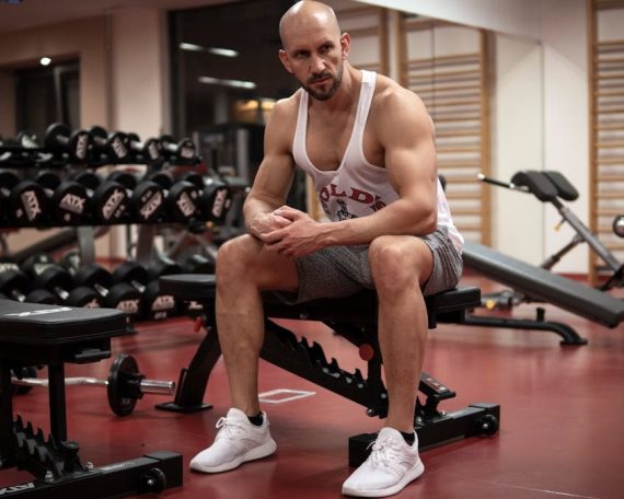 Strong, determined man in white tank top sitting in gym, showcasing fitness and strength.